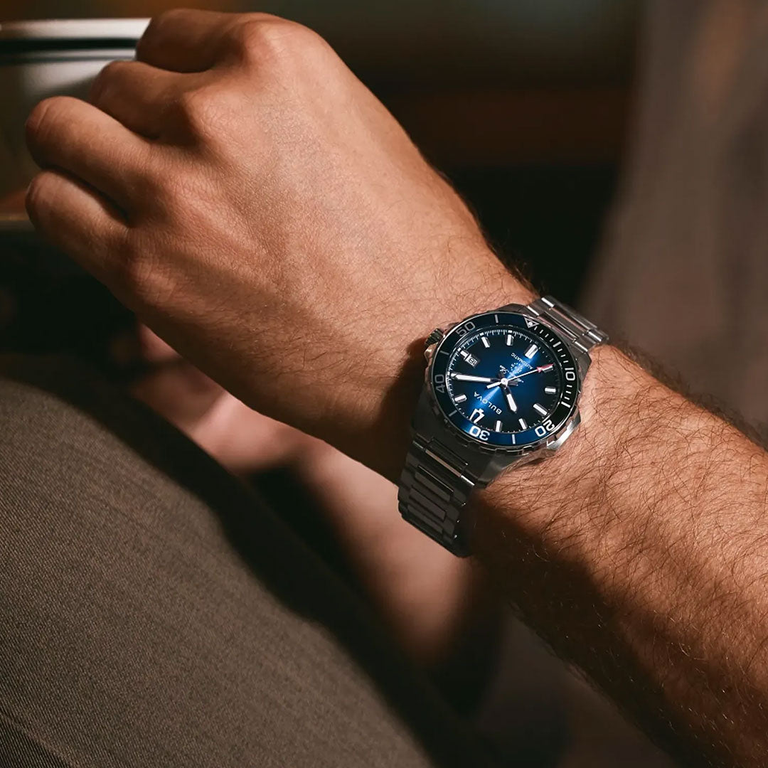 A person’s wrist wearing a Bulova Marine Star Series B Automatic 41mm Watch with a blue dial and silver band, holding a cup. The Bulova timepiece stands out in a relaxed, casual setting.