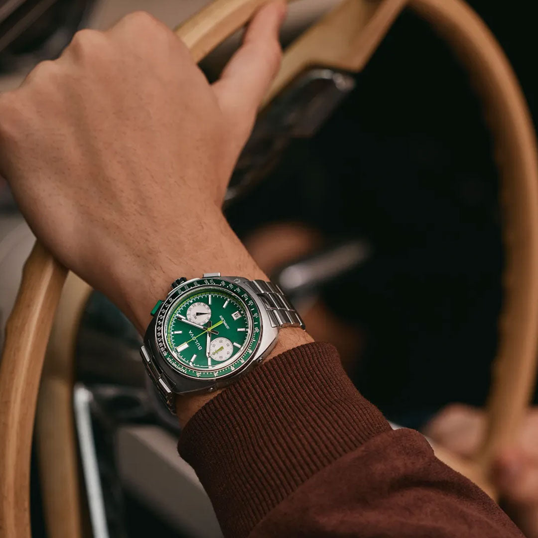 Wearing a brown long-sleeve shirt, a person grips a wooden steering wheel while driving, sporting the Bulova Racer Quartz 42mm Watch—a stainless steel timepiece with a green dial and two white subdials by Bulova.