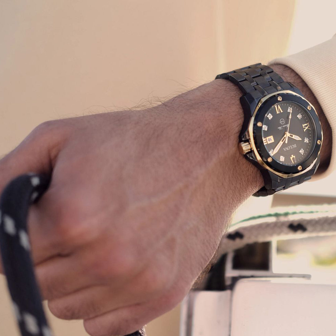 A close-up of a person's hand holding a black rope showcases the sleek sport luxury design of the Bulova Marine Star Quartz 44mm Watch. This black wristwatch from Bulova's Marine Star Collection features gold accents and Roman numerals on its black ion-plated stainless steel strap, elegantly standing out against the blurred background.