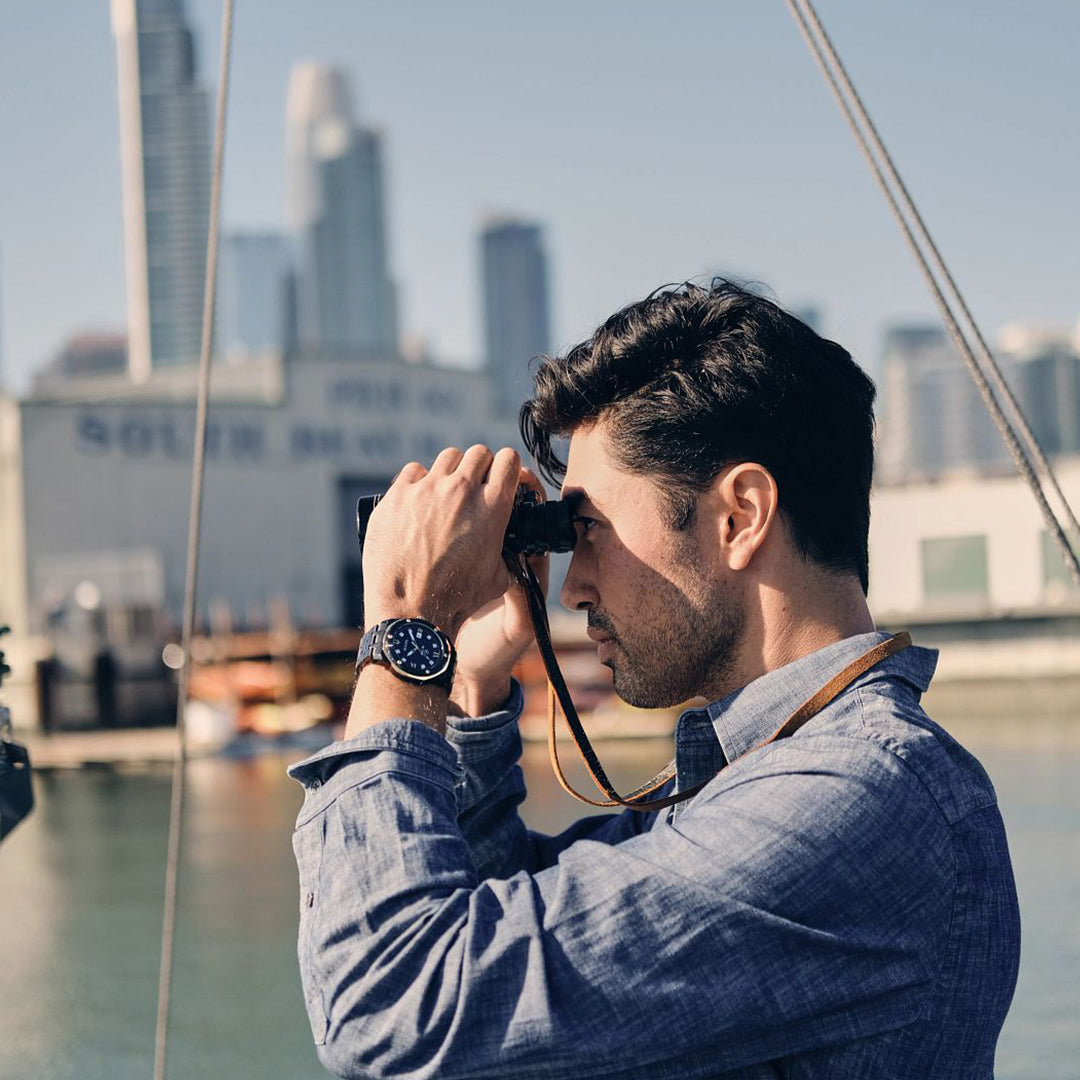 A person dressed in a blue shirt is near a waterfront, peering through binoculars while featuring the sport luxury design of the Bulova Marine Star Quartz 44mm Watch. The industrial skyline and clear sky enhance the appeal of their black ion-plated stainless steel timepiece by Bulova.