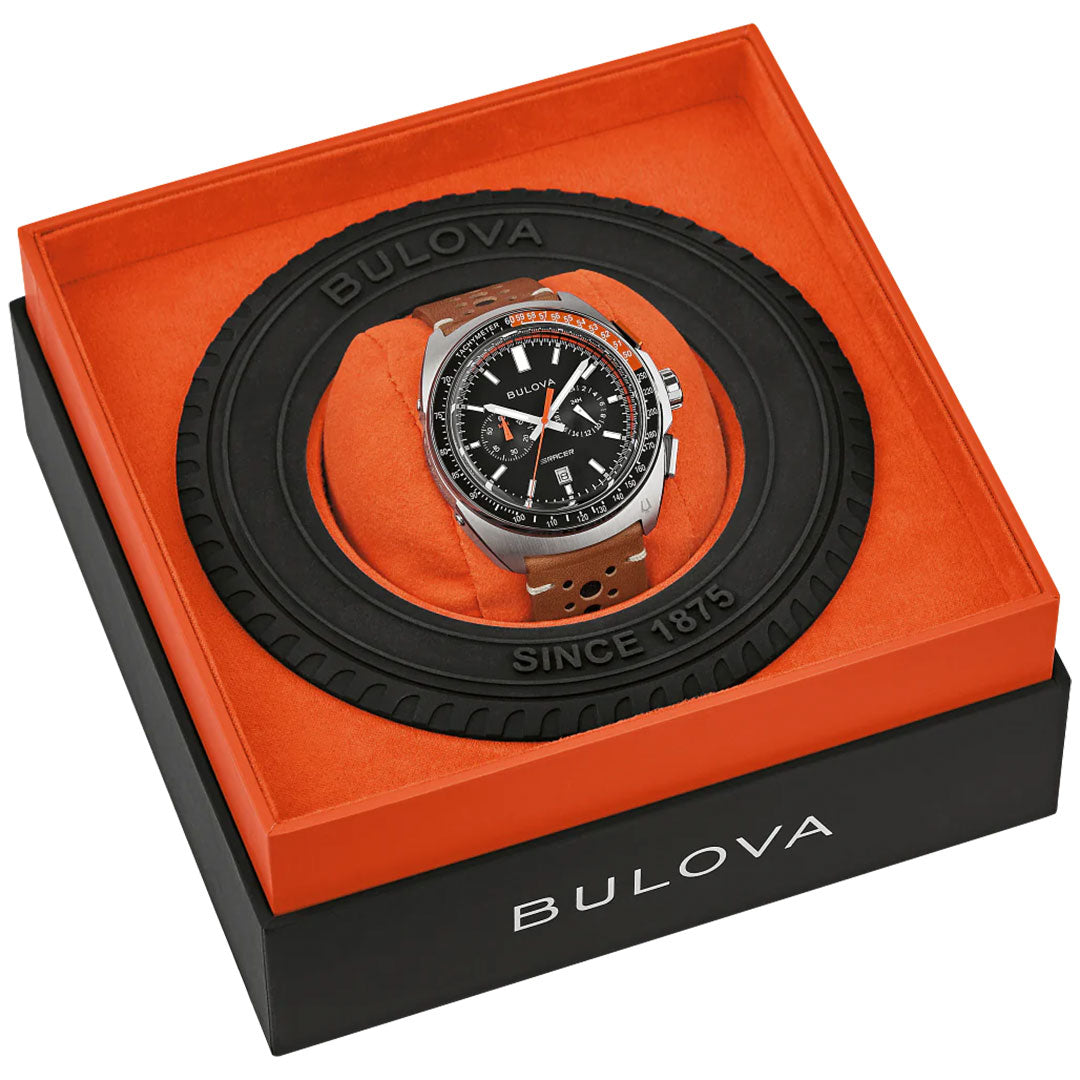 The Bulova Racer Quartz 42mm Watch is shown in an orange and black box with a round, tire-inspired frame. The brown leather strap and stainless steel case are visible, with the Bulova brand name on both the box lid and frame.