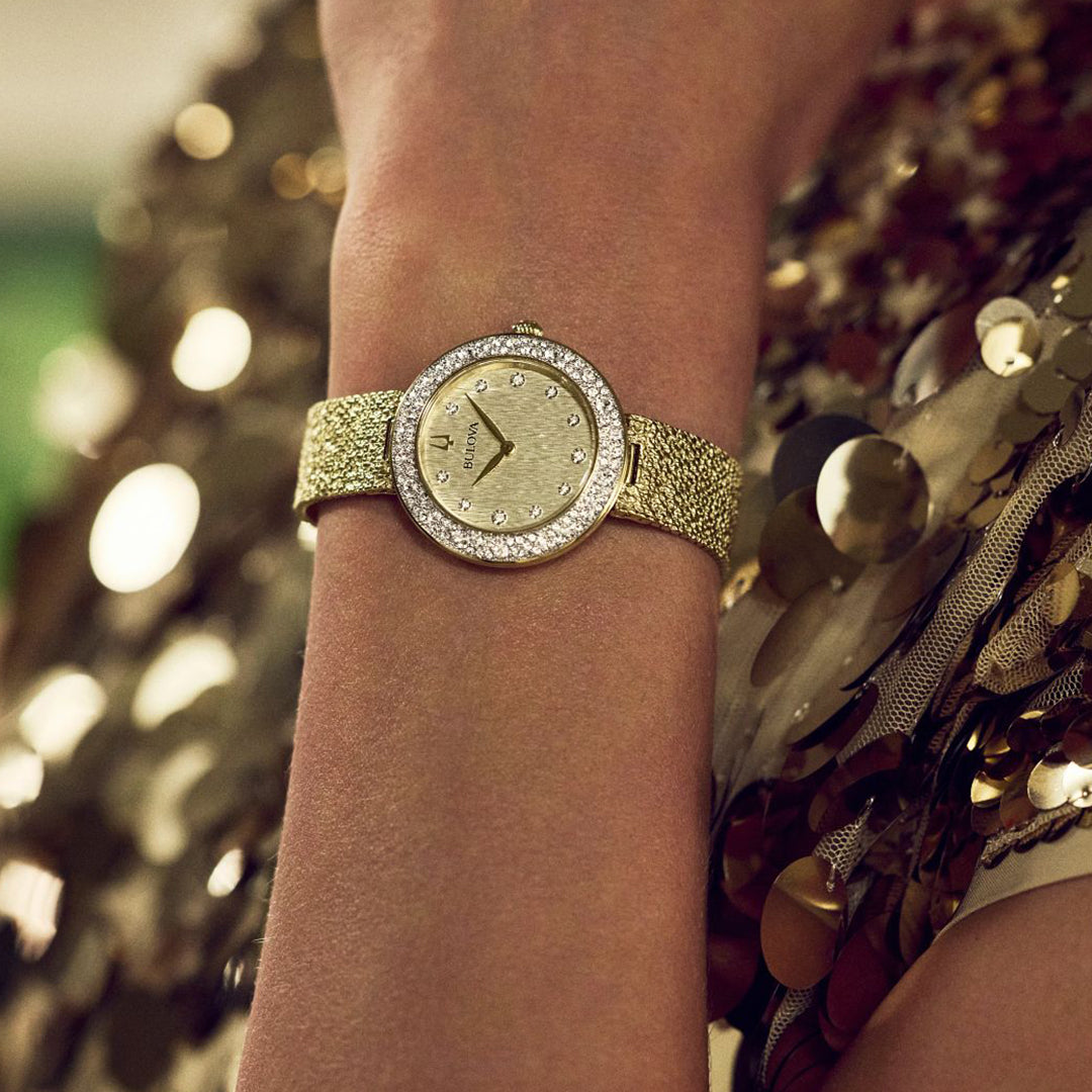 Close-up of the Bulova Crystal Quartz 32mm Watch, featuring a gold-textured strap and diamond-studded bezel. The champagne dial with minimalist hands rests on shimmering fabric with golden sequins, enhancing its luxury.