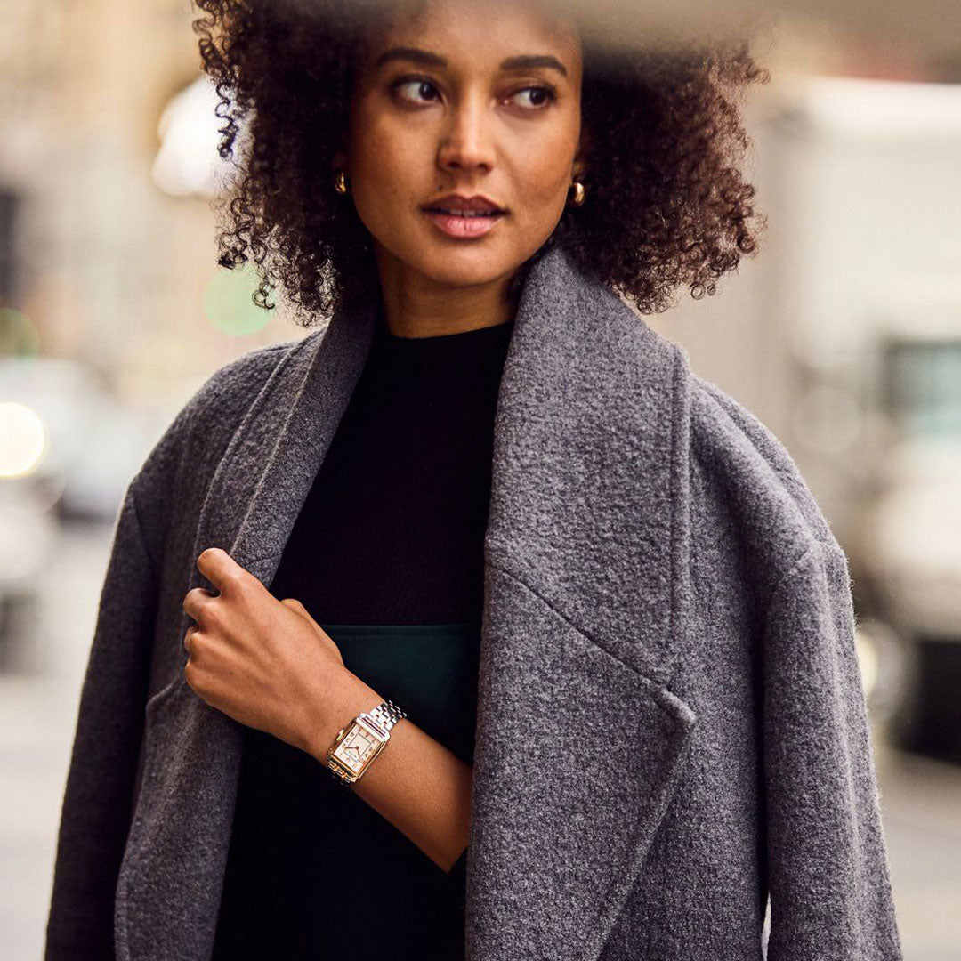 A woman with curly hair in a gray coat wears the Bulova Sutton Quartz 25mm Watch, standing outdoors in an urban scene. A softly blurred background highlights her as the main subject, drawing attention to her and the Bulova watch.
