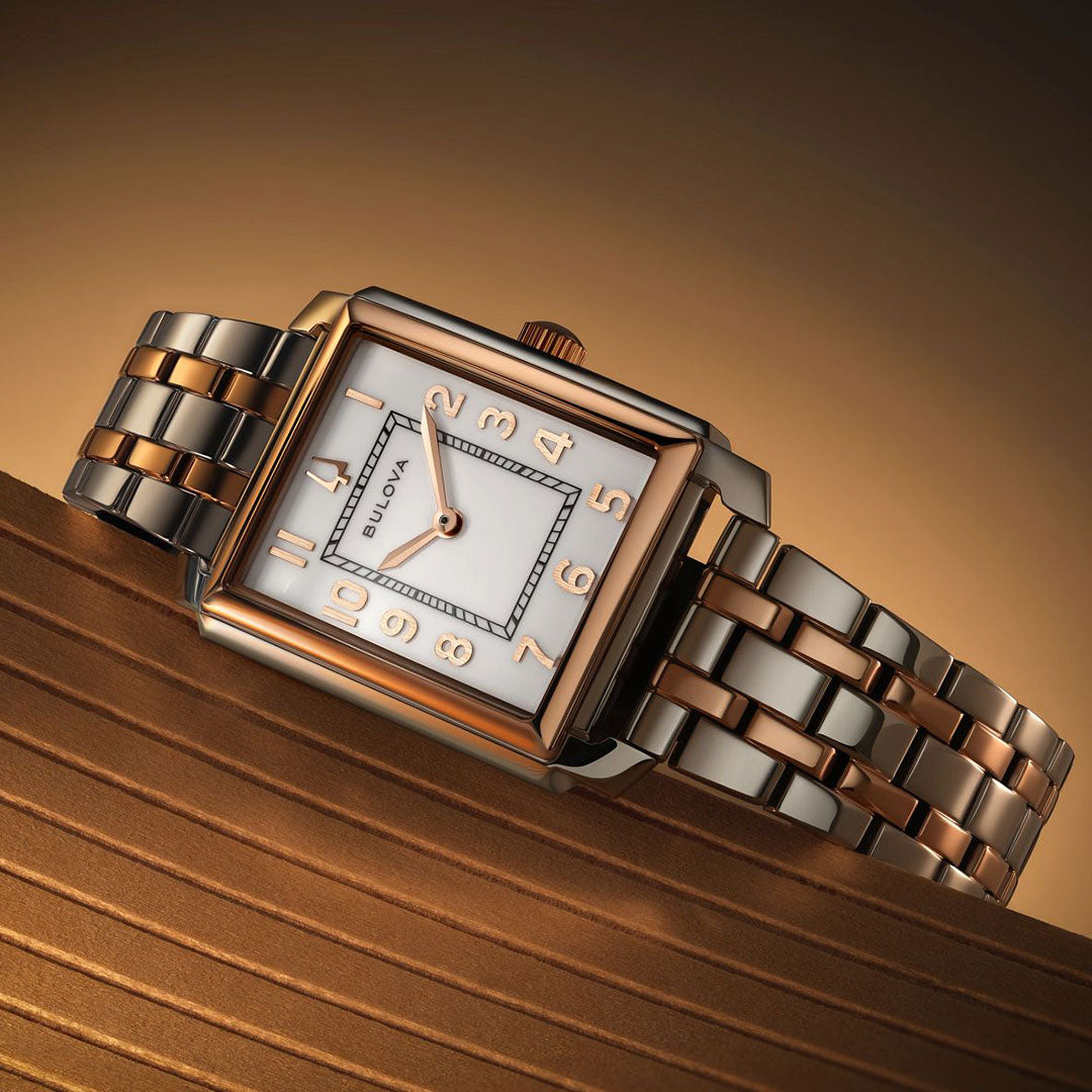 The Bulova Sutton Quartz 25mm women's watch features a rectangular two-tone case, white dial with gold-tone numerals, and a silver and rose gold stainless steel link band, shown against a warm brown gradient background.
