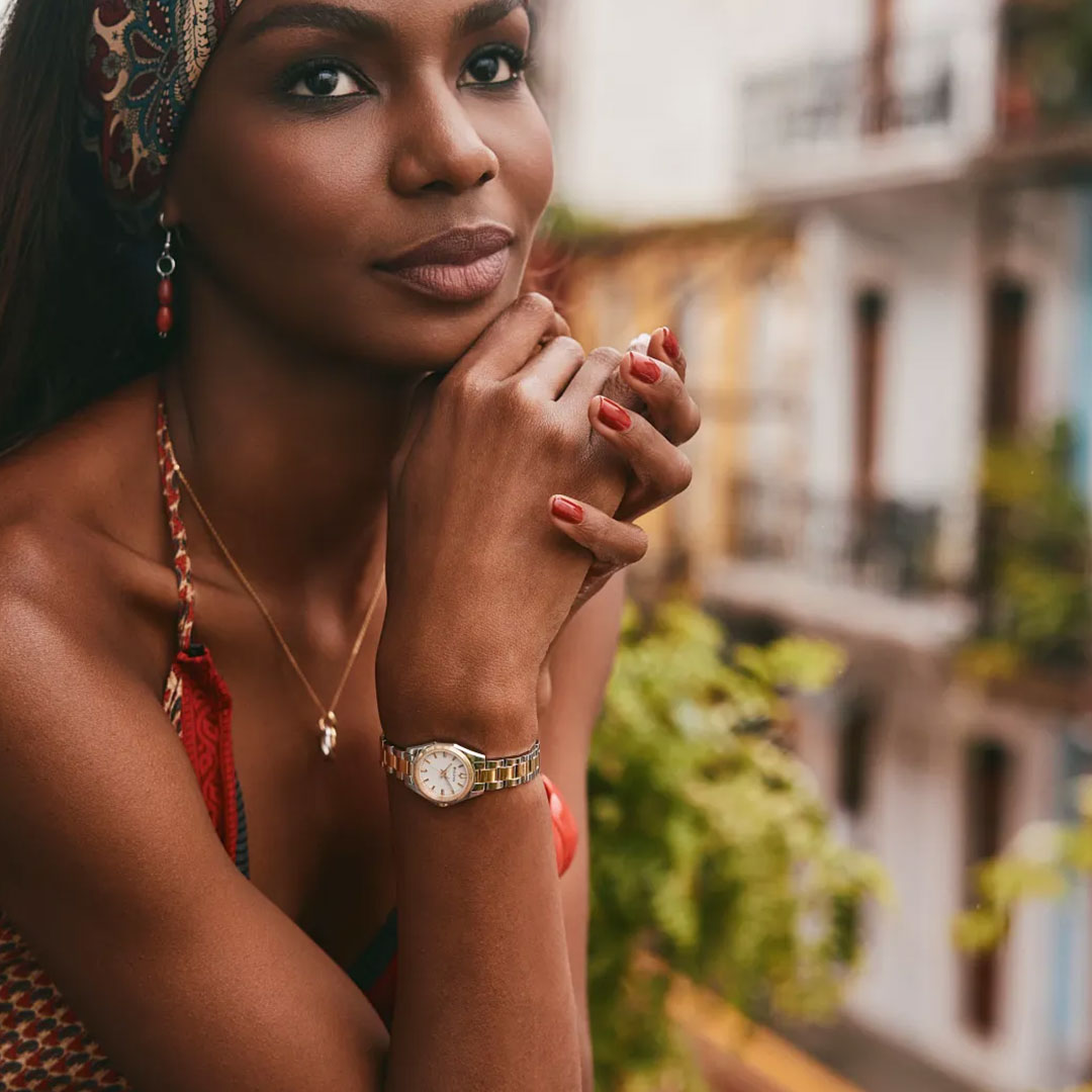 A woman with a headscarf and red nails rests her chin on her hands, highlighting the Bulova Surveyor Quartz 22.7mm Watch by Bulova, as she stands on a balcony with plants and colorful buildings behind her.