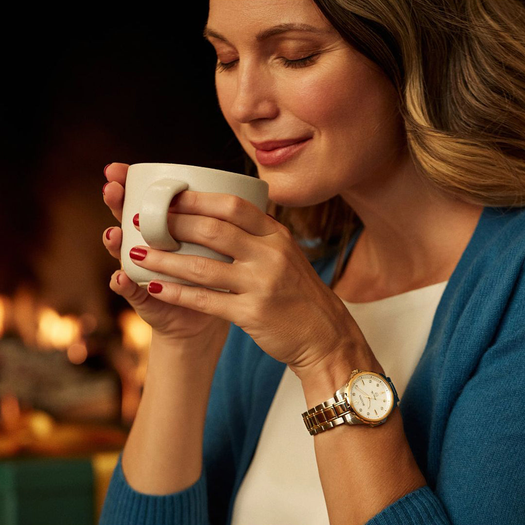 A woman in a blue sweater holds a white mug, smiling with her eyes closed. She wears the Bulova Marine Star Quartz 36mm Watch, featuring a mother-of-pearl dial, and has red nails. A fireplace glows warmly in the background, creating a cozy atmosphere reminiscent of the elegance of this Bulova ladies' timepiece.