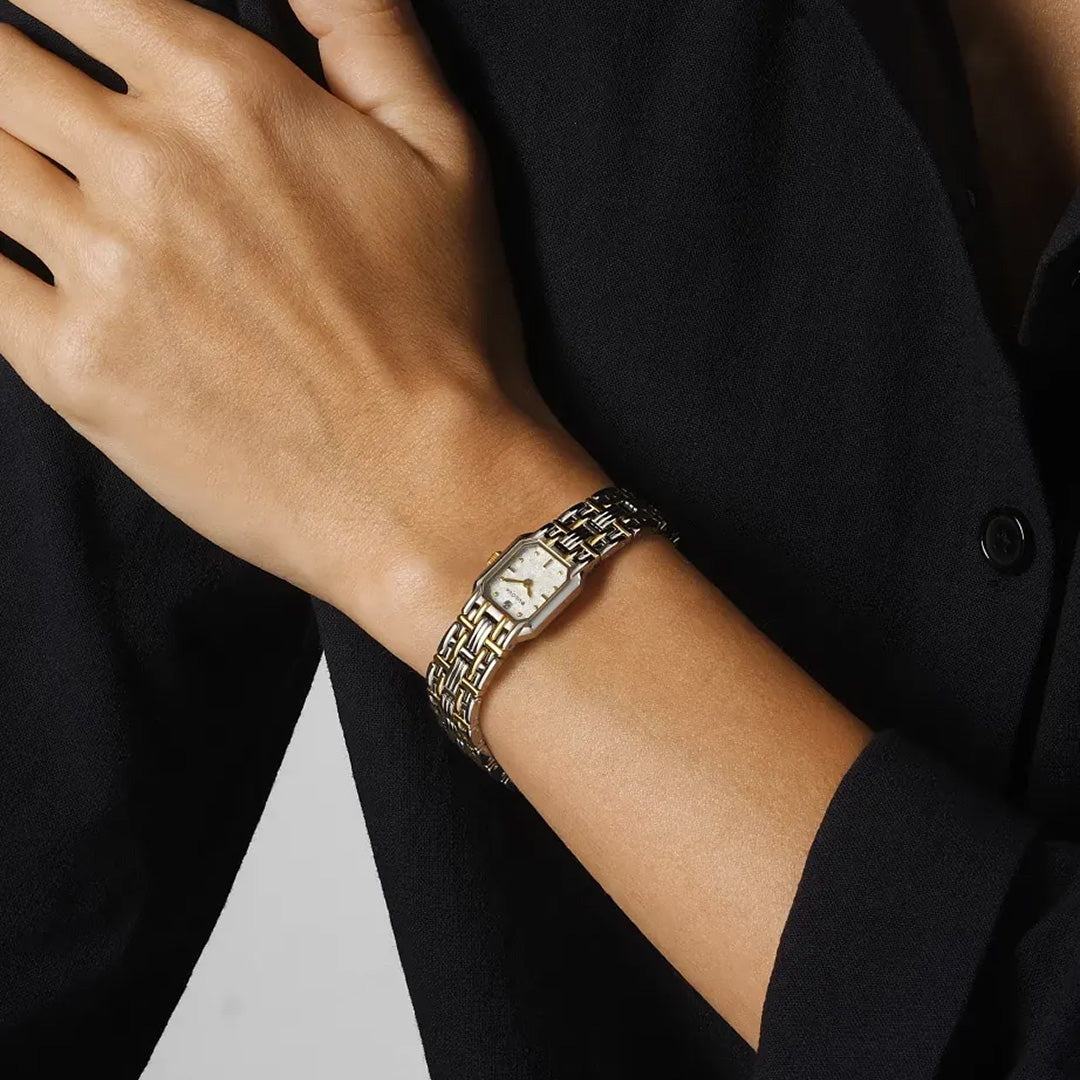 A person wearing a black long-sleeve shirt displays the Bulova Goddess of Time Sophia Quartz 16mm Watch on their left wrist—a gold wristwatch by Bulova featuring a rectangular face and patterned metal band that echoes the elegant neo-vintage style of a classic 1993 Bulova recreation.