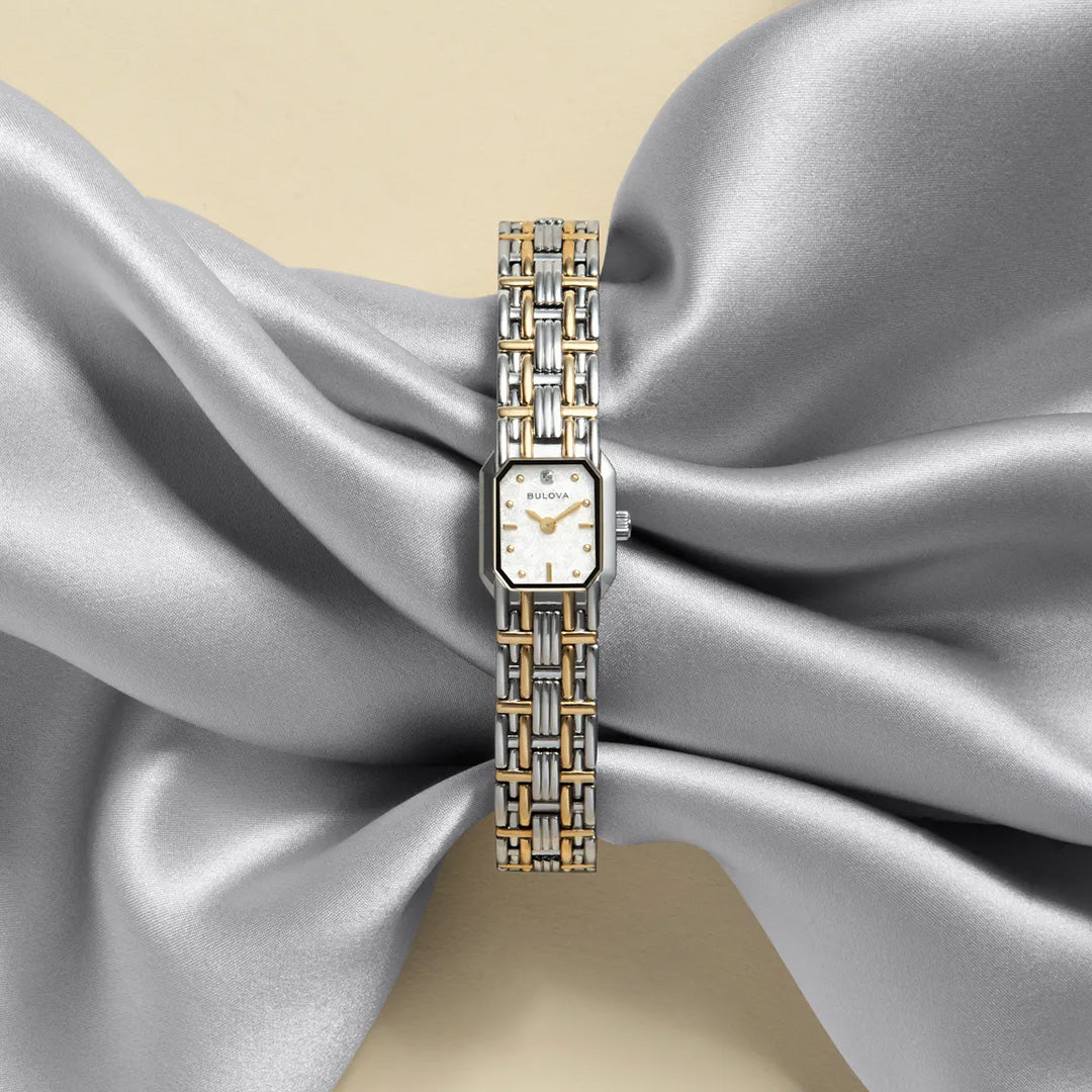 A Bulova Goddess of Time Sophia Quartz 16mm Watch, in silver and gold tones, rests elegantly atop a metallic silver fabric folded gracefully like a bow, set against a soft beige background.