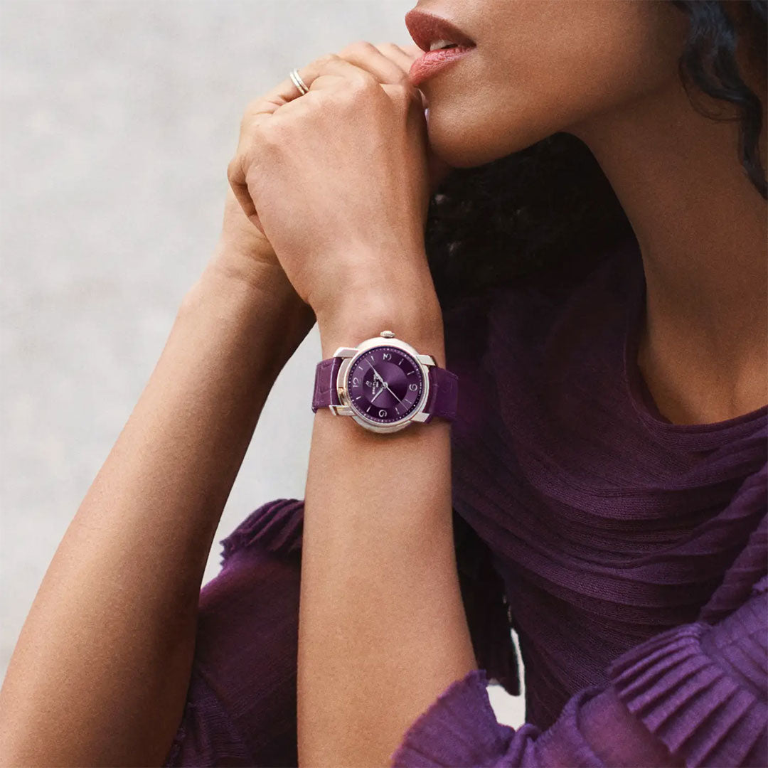 A woman wearing a textured purple top rests her chin on her hand, showcasing a Breitling Lady Premier Automatic 36 watch with a purple strap and matching purple dial.