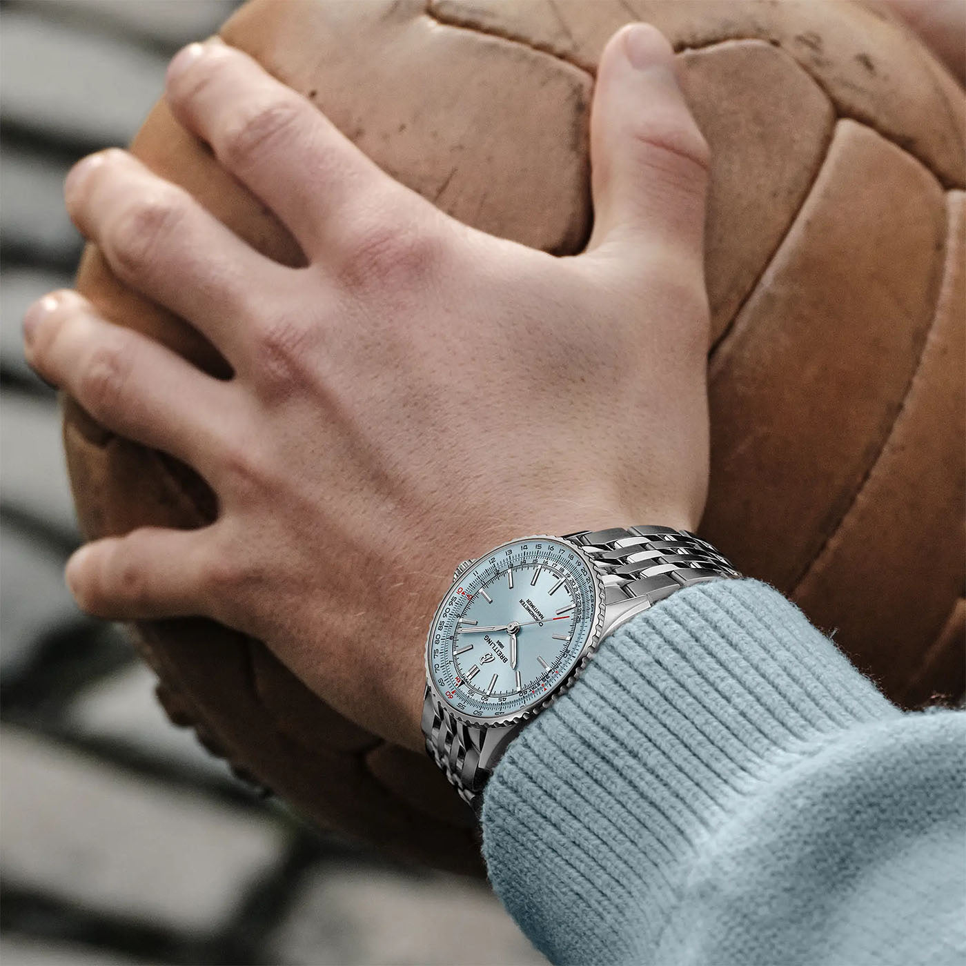 A hand adorned with a BREITLING Navitimer Automatic 41, showcasing a light blue face, grasps a vintage leather soccer ball. The person is dressed in a light blue sweater, all set against the charming backdrop of cobblestone paving.