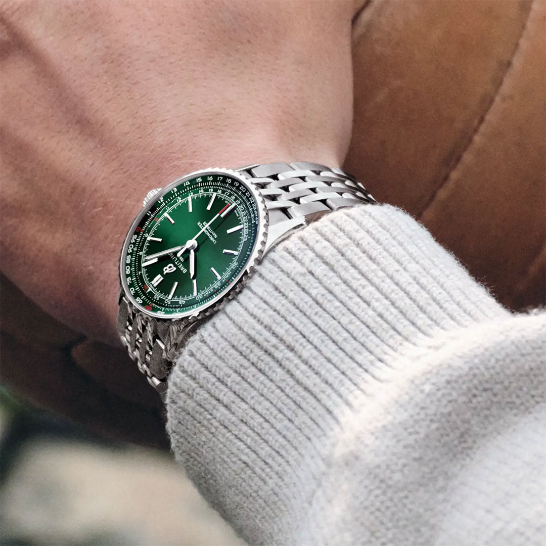 Wearing a light sweater, a person showcases the BREITLING Navitimer Automatic 41 by Breitling—a luxury pilot watch with a green dial and metal strap—while holding a brown leather object, highlighting its elegant GMT-inspired style.