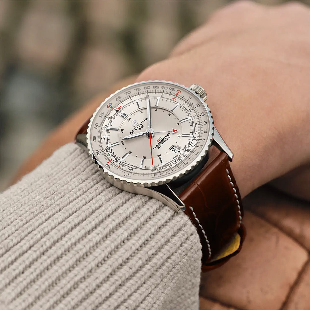 A close-up shows a wrist wearing the Breitling Navitimer Automatic GMT 41 with a silver dial and brown leather strap, styled with a light knit sweater for a refined, elegant look.