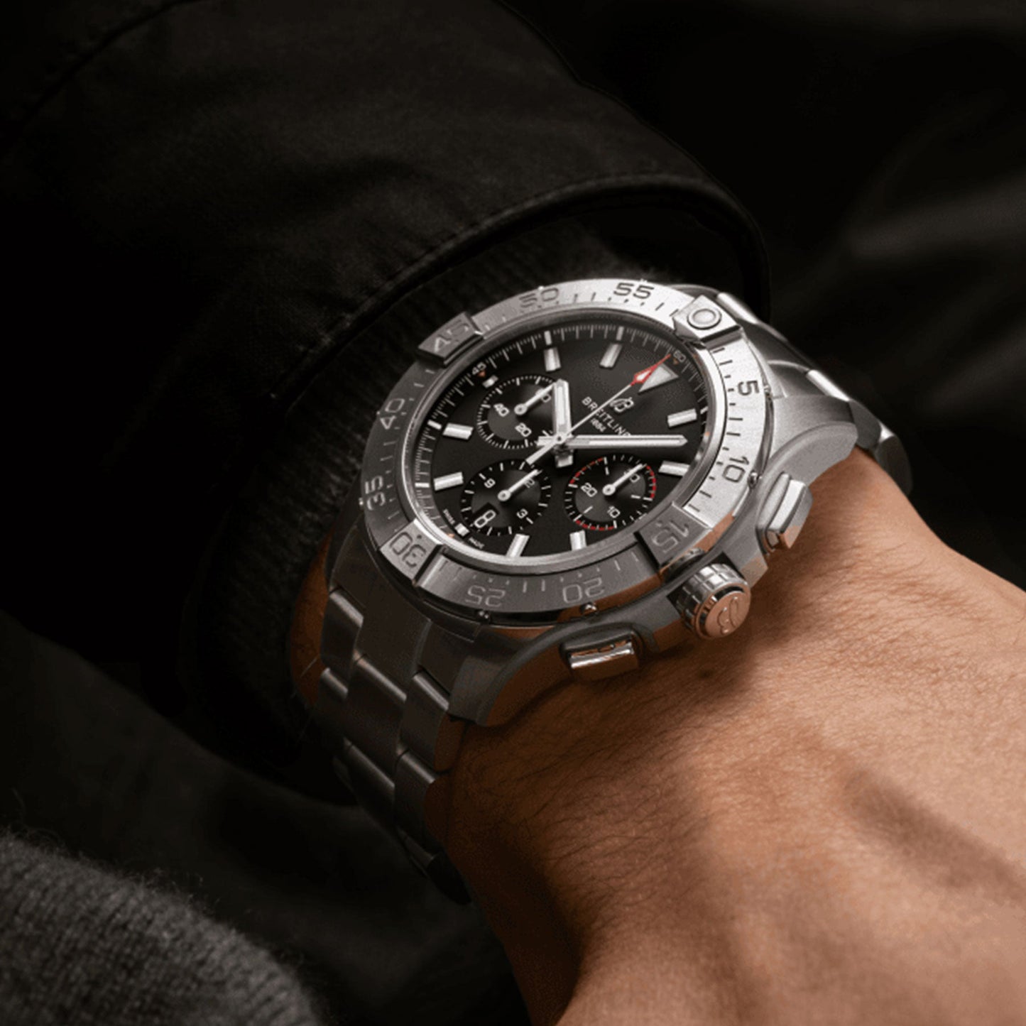 Close-up of a person's wrist wearing the Breitling Avenger B01 Chronograph 44, featuring a black face and multiple dials. The dark background emphasizes the watch's intricate details and highlights its chronograph movement.