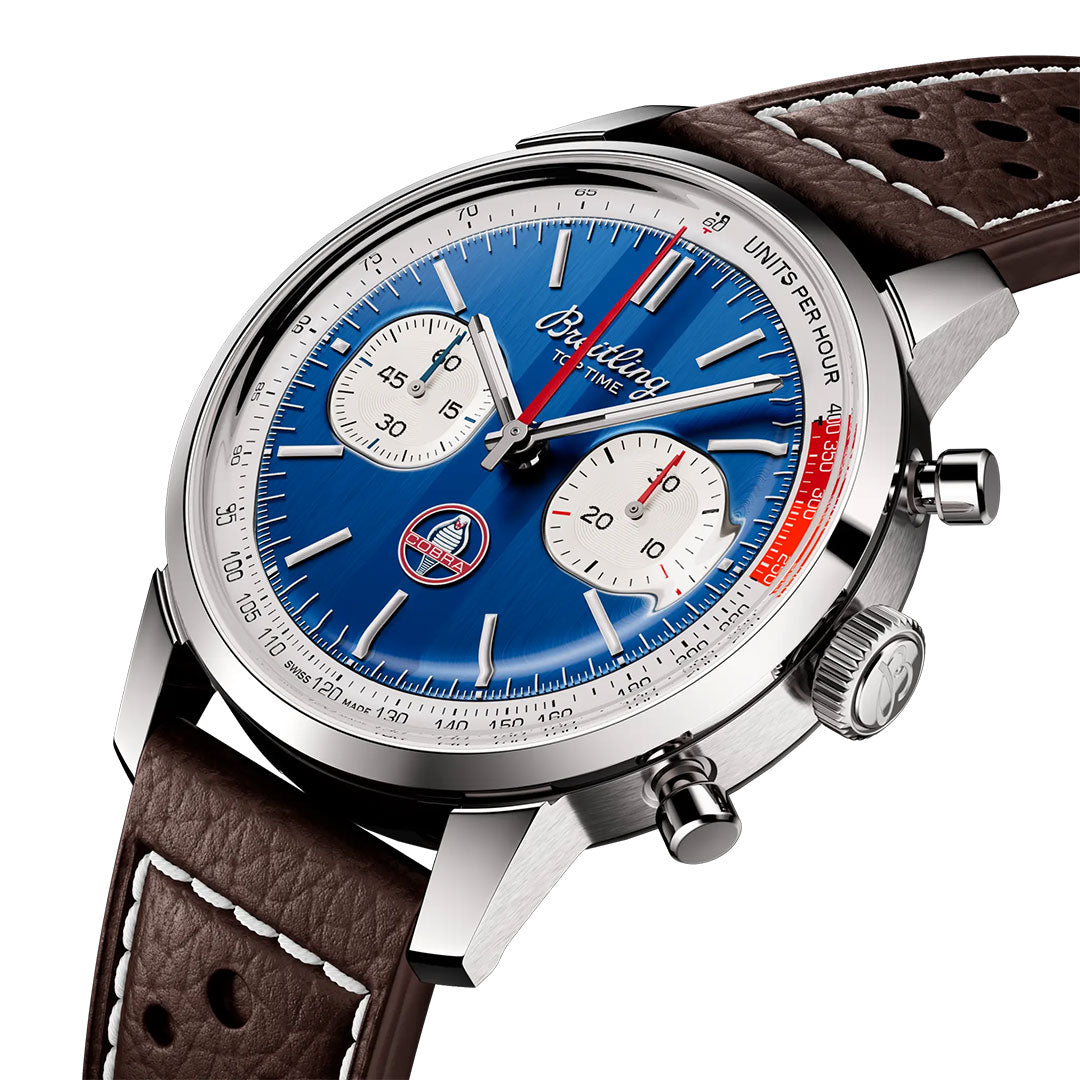 The Breitling Top Time B01 Shelby Cobra is a luxury chronograph with a blue dial, white subdials, silver indices, and brown perforated leather strap. It features chrono pushers, tachymeter scale, and detailed strap stitching inspired by the Shelby Cobra.