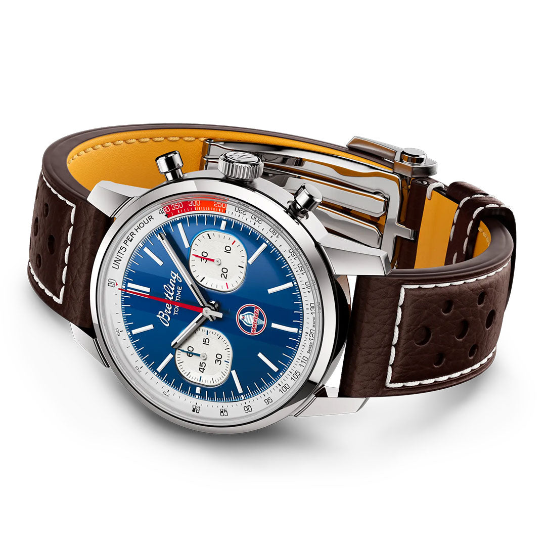 The BREITLING Top Time B01 Shelby Cobra features a blue dial with two white subdials, red and white accents, silver casing, and a brown perforated leather strap with white stitching.