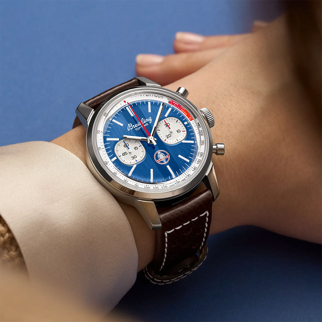 A person wears the Breitling Top Time B01 Shelby Cobra chronograph with a blue dial, two white subdials, red second hand, and brown leather strap, set against a light blue background.
