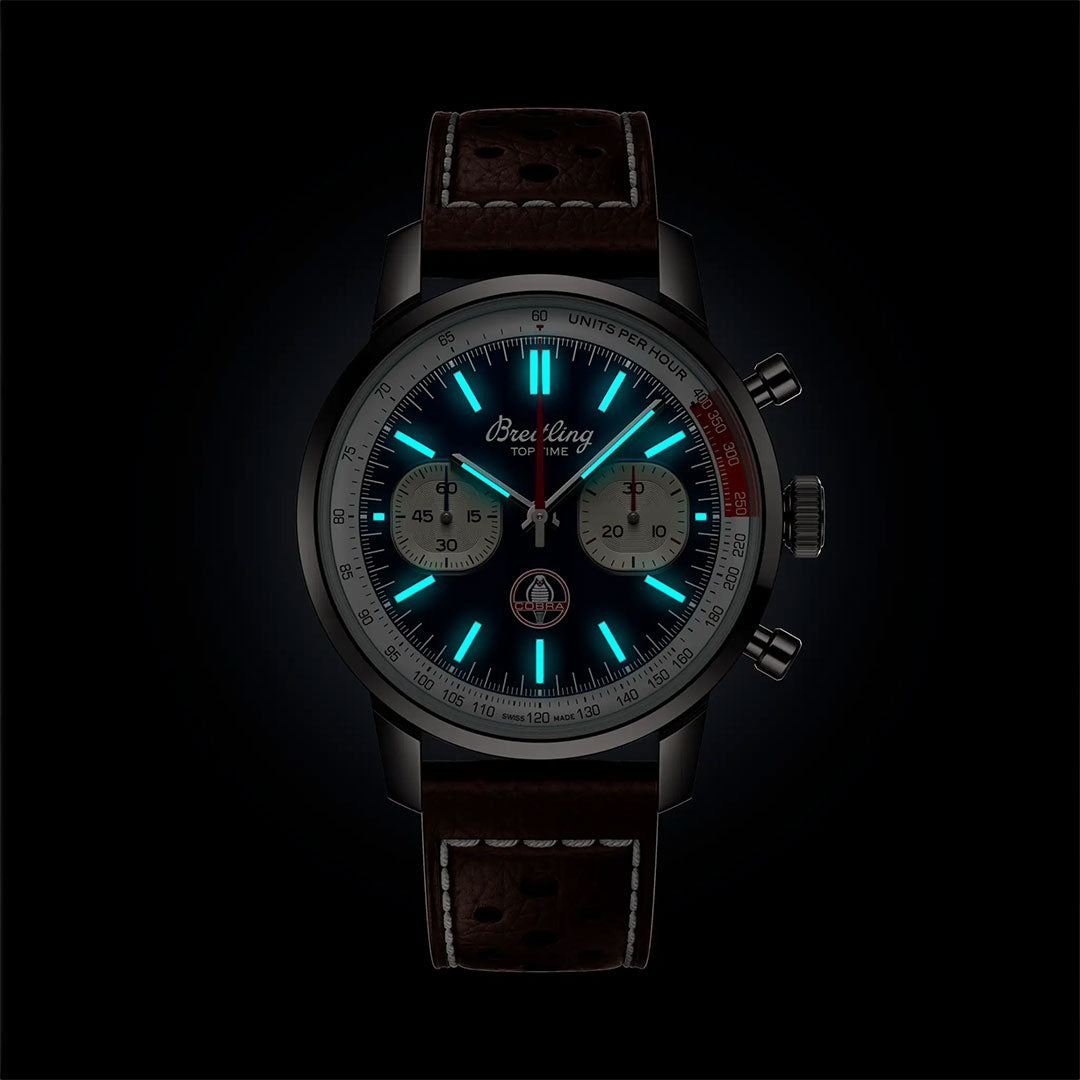 The BREITLING Top Time B01 Shelby Cobra by Breitling features a dark brown leather strap, glowing dial with blue luminescent hour markers, two subdials, and the model name on the face, shown against a bold black background.