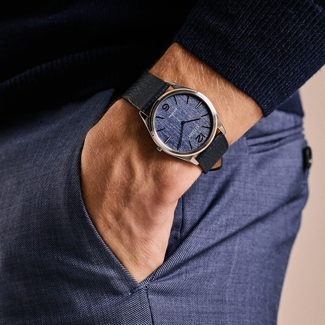 A person wearing a navy sweater and blue trousers has their hand in a pocket, showcasing the CITIZEN Suratto Eco-Drive 38.4mm Watch with a leather strap. The silver-tone stainless steel bezel frames the textured face with minimal hour markers, highlighting its stylish design.