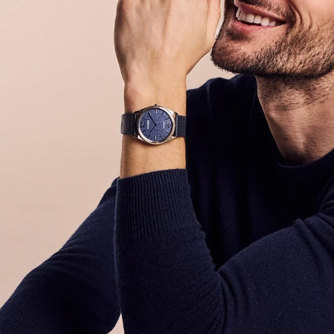 A person in a dark sweater smiles, resting their chin on their hand, wearing a CITIZEN Suratto Eco-Drive 38.4mm Watch, which has a sleek navy blue face and black strap.