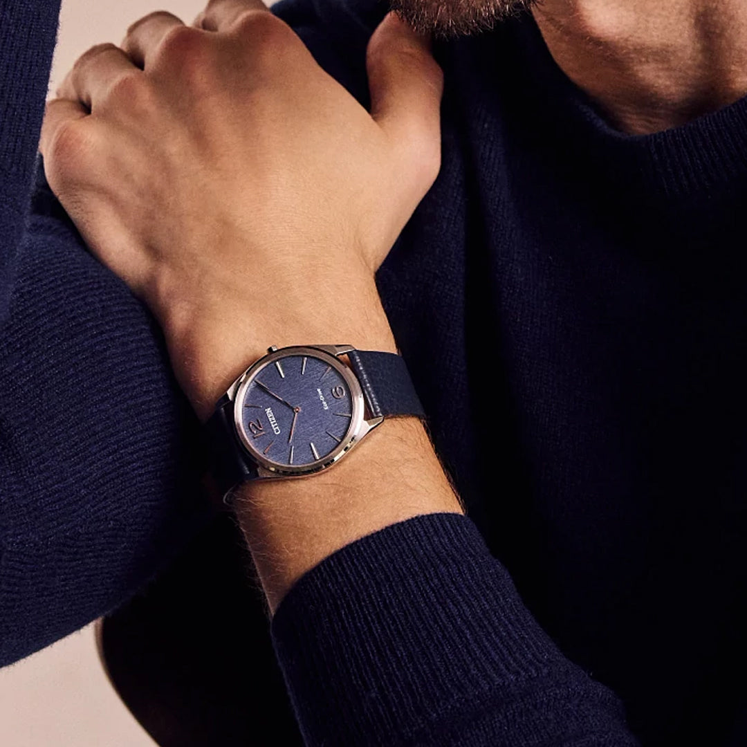 A person in a dark blue sweater showcases the CITIZEN Suratto Eco-Drive 38.4mm watch with a blue face, dark strap, and silver hour markers. Its minimalist design and water resistance are highlighted as their arm is crossed over their chest.