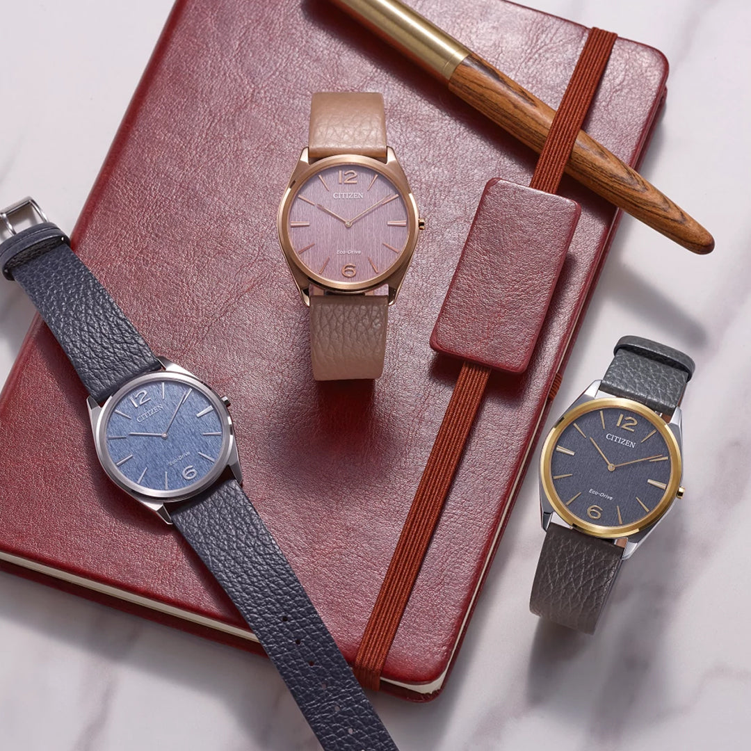 Three CITIZEN Suratto Eco-Drive 38.4mm watches with textured leather straps are displayed on a red notebook. Featuring rose gold, silver, and gold-toned cases with pink, blue, and grey dials, these elegant timepieces include Eco-Drive technology. A brown pen is nearby.