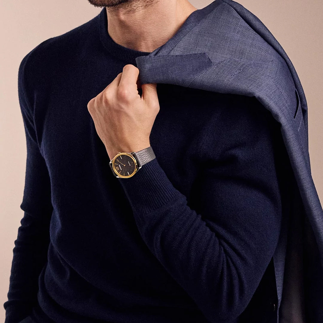 A person dons a dark sweater, draping a blue blazer over their shoulder while wearing the elegant CITIZEN Suratto Eco-Drive 38.4mm Watch, featuring water-resistant technology, a gold case, and a dark face. The background is neutral.