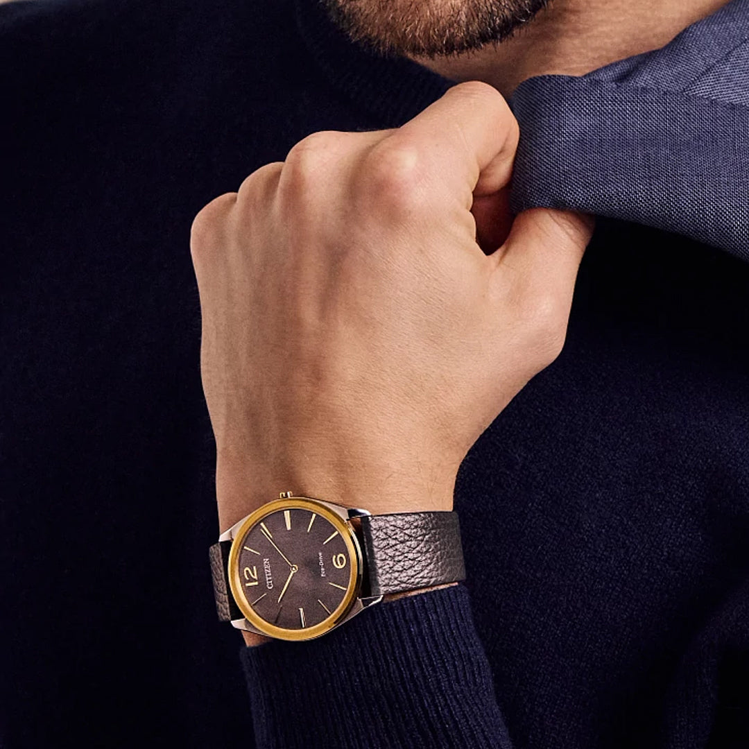 A person in a dark sweater adjusts a light blue collar. On their wrist, the CITIZEN Suratto Eco-Drive 38.4mm Watch shines with its gold-colored rim, dark face, and leather strap, showcasing minimalist design and water resistance.