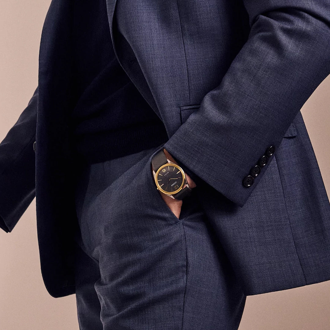 A person in a dark blue suit with their hand in their pocket wears the CITIZEN Suratto Eco-Drive 38.4mm watch, featuring a sleek two-tone stainless steel design. The black face with a gold rim enhances its stylish and professional look.