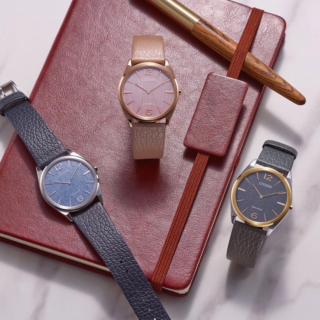 A CITIZEN Suratto Eco-Drive 38.4mm watch, available with blue, pink, or grey dials and leather straps, is stylishly displayed on a red leather notebook beside a gold and wood pen. Combining classic design with innovation, these watches exemplify elegance and Eco-Drive technology.