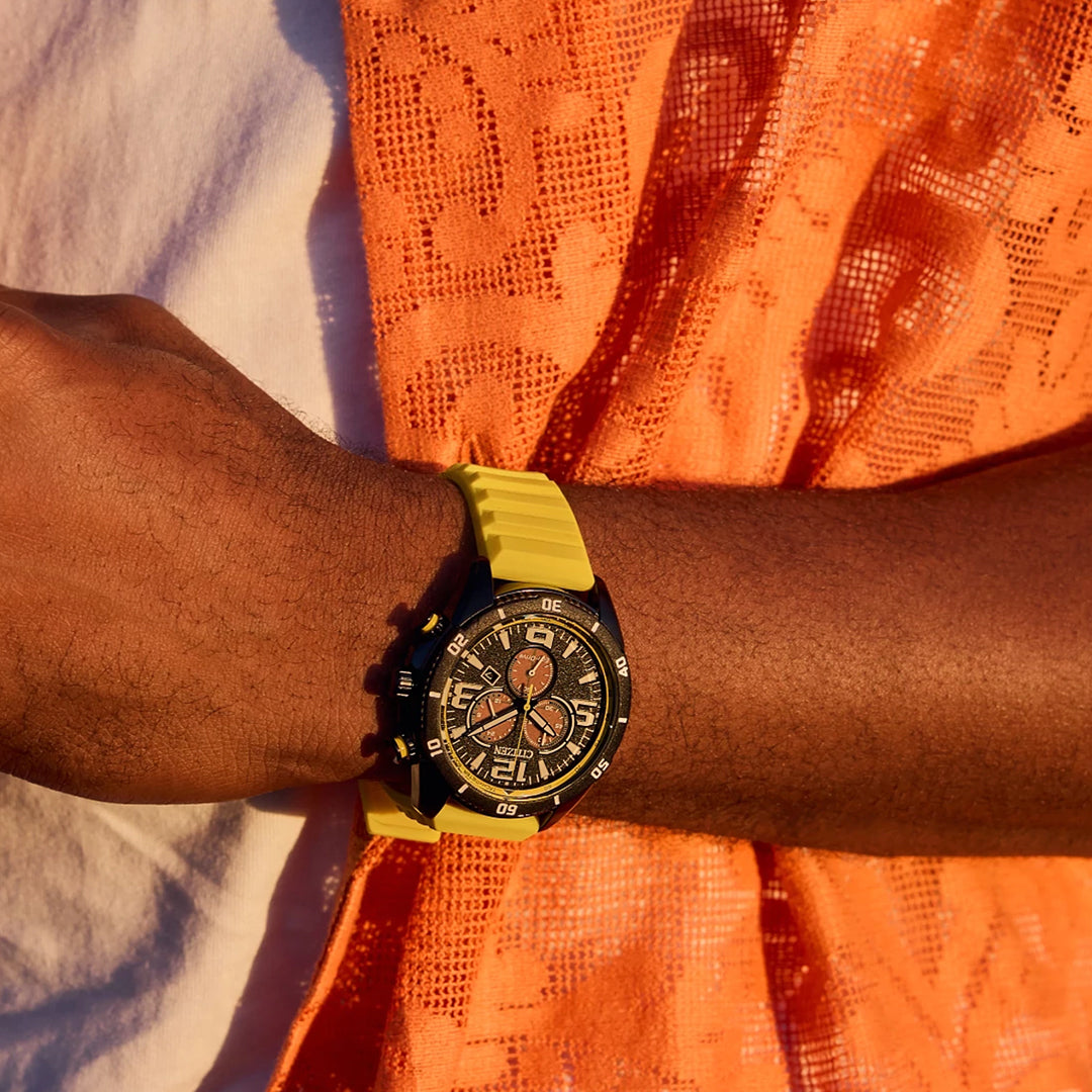 A person is seen wearing a CITIZEN Brycen Eco-Drive 44.8mm watch, featuring a detailed dial and black-yellow band. They hold orange lace fabric against white clothing, with warm lighting highlighting the colors.