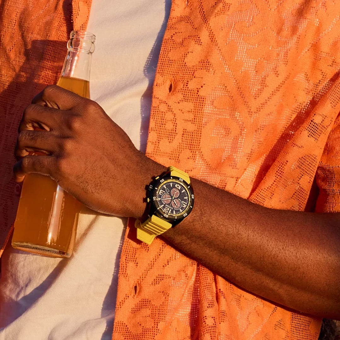 A person in an orange lace shirt holds an orange soda, wearing a CITIZEN Brycen Eco-Drive 44.8mm Watch on their wrist. Sunlight casts playful shadows, reflecting the vibrant interplay of light and fashion.