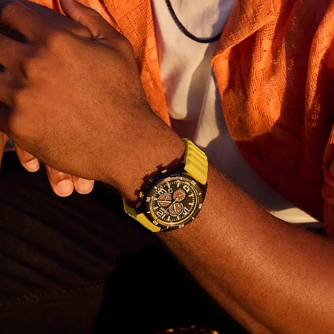 A person in a vibrant orange shirt sits with hands clasped, wearing the CITIZEN Brycen Eco-Drive 44.8mm Watch. The watch features a yellow strap and chronograph details, complementing the softly lit background that creates a warm and relaxed atmosphere.