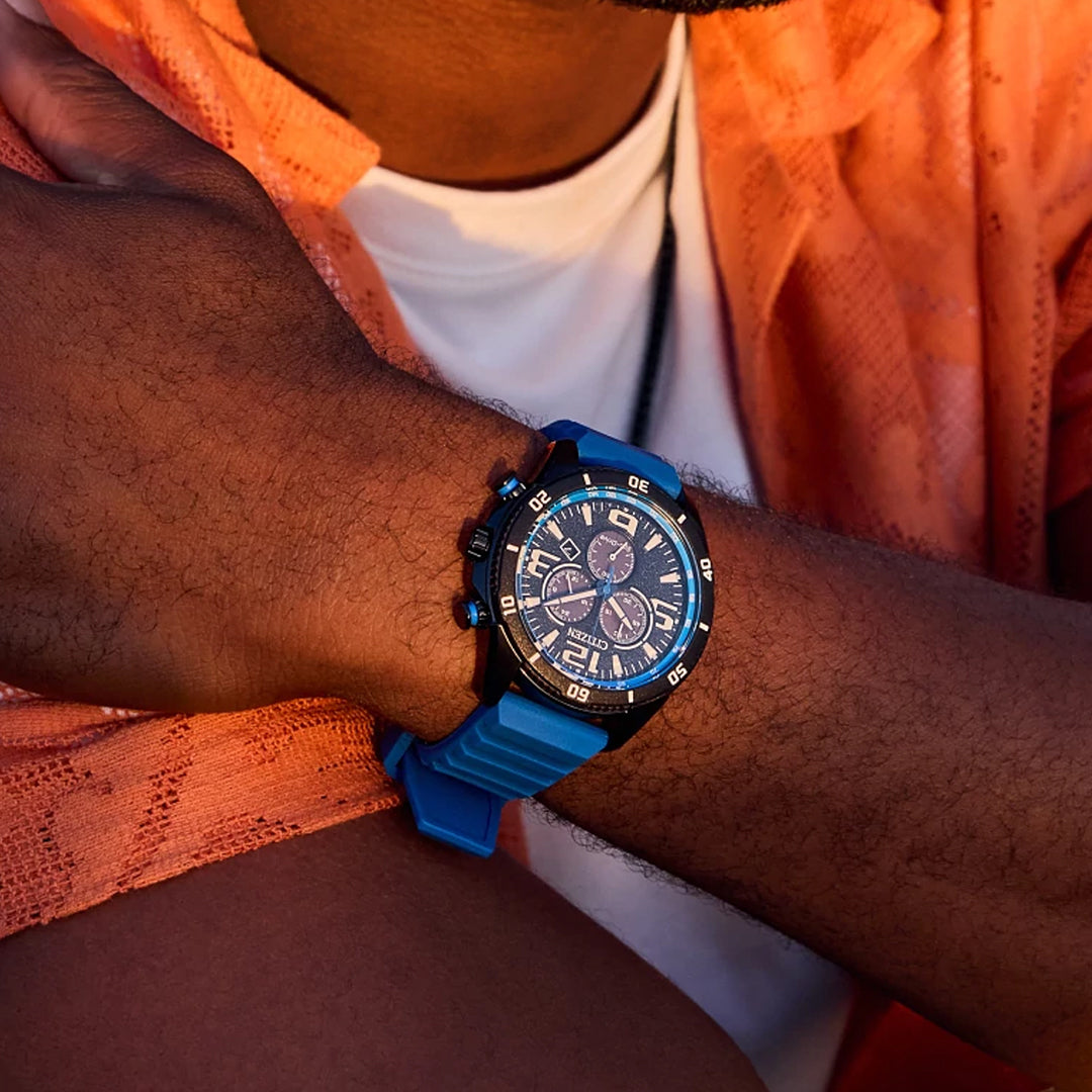 A person in an orange shirt and white tee is showcasing a CITIZEN Brycen Eco-Drive 44.8mm Watch on their wrist, which features a detailed face with multiple dials and a blue strap, set against a blurred outdoor background.