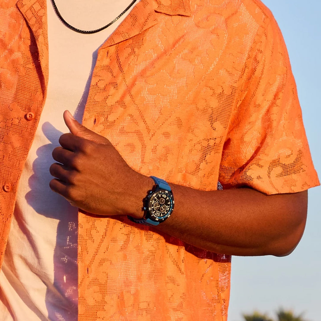 A person in a patterned orange shirt over a white tee is shown from the neck down, outdoors on a sunny day. They wear a CITIZEN Brycen Eco-Drive 44.8mm watch on their left wrist and grasp their shirt with their right hand, illustrating the perfect mix of style and functionality.