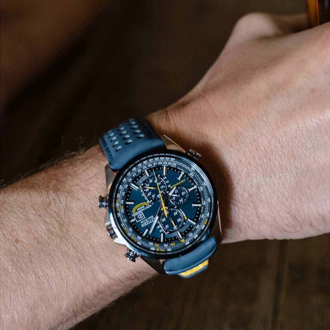 Close-up of a CITIZEN Blue Angels World Chronograph AT Eco-Drive 43mm watch with a dark blue face, multiple dials, yellow accents, radio-controlled features, and a perforated blue strap. It's worn on a person's wrist against a wooden background.