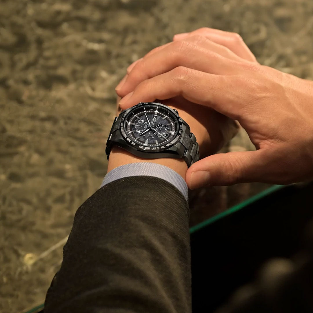 A person in a dark suit checks the time on a CITIZEN Attesa Hakuto-R Atomic Time Keeping Super Titanium Limited Edition Eco-Drive 42mm watch, standing by a marble-patterned surface.
