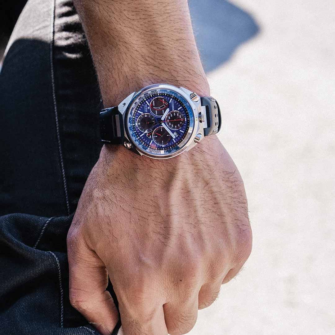 Close-up of a person's hand wearing the CITIZEN Promaster Tsuno Chrono Racer E210 Eco-Drive 43.3mm Limited Edition watch, which features a silver case, blue dial with multiple subdials, and a black strap. The watch exudes a sporty, modern design. The background is slightly blurred, emphasizing the watch as the main focus.