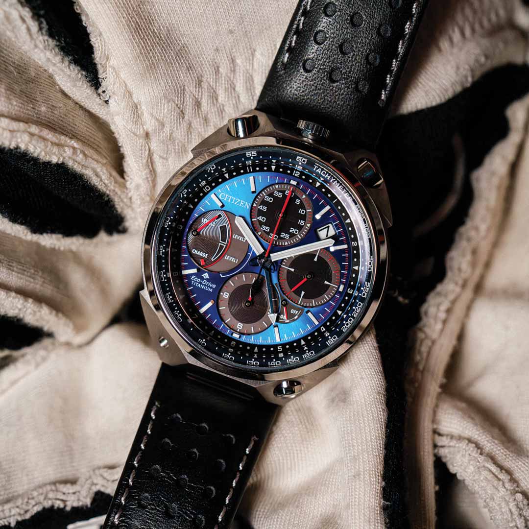 A close-up of the CITIZEN Promaster Tsuno Chrono Racer E210 Eco-Drive 43.3mm Watch Limited Edition reveals its intricately designed chronograph with multiple dials and a black leather strap. The watch face features detailed red and blue accents, set against a textured black and white surface.