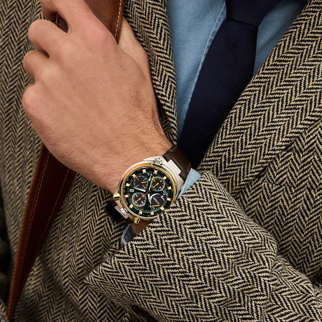 A person in a herringbone-patterned jacket, blue shirt, and dark tie holds a strap. On their wrist is the CITIZEN Promaster Tsunoc Chrono Racer Limited Edition Eco-Drive 44.5mm Watch, distinguished by its brown leather strap and black-and-gold face—renowned for CITIZEN’s precision craftsmanship and innovative Eco-Drive technology.
