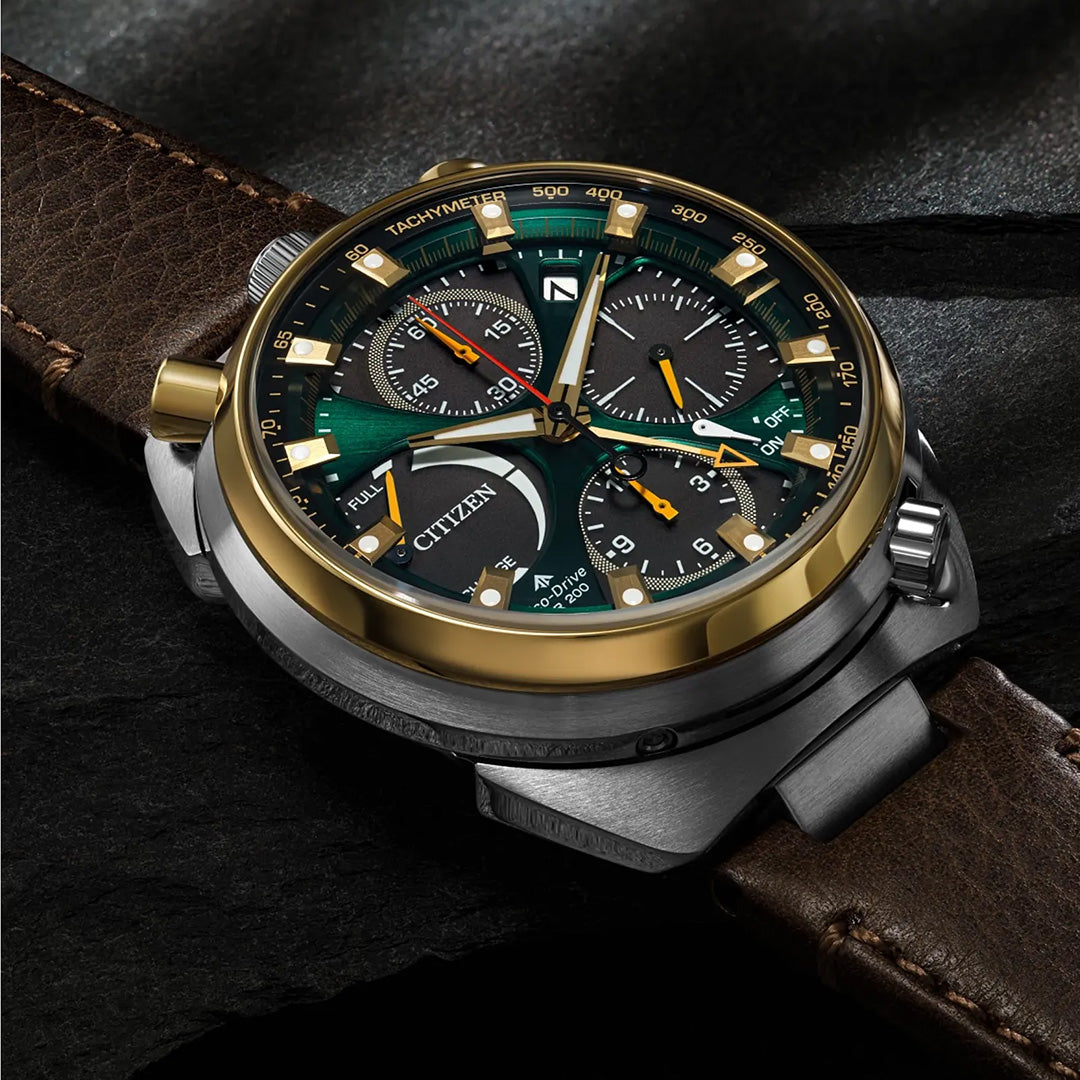 The CITIZEN Promaster Tsunoc Chrono Racer Limited Edition Eco-Drive 44.5mm Watch by CITIZEN, with its striking green and gold bezel, brown leather strap, multi-dial face, and silver case, showcases innovative Eco-Drive technology as it rests elegantly on a dark surface.
