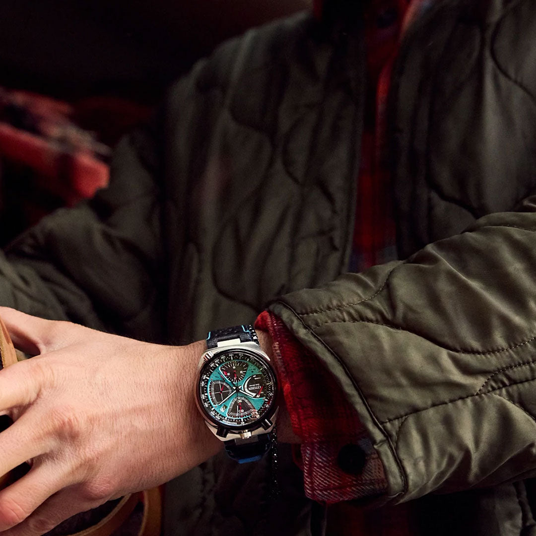 Wearing a green quilted jacket and red plaid shirt, a person displays their wrist adorned with the CITIZEN Promaster Tsuno Chrono Racer Limited Edition Eco-Drive 44.5mm Watch, showcasing its modern multi-colored face and black strap.