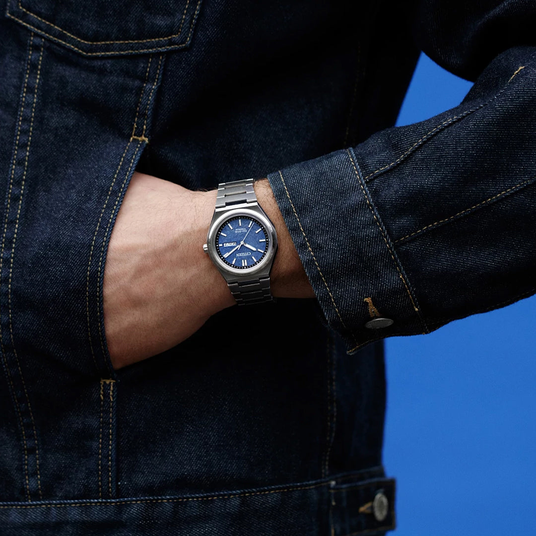 A person in a dark denim jacket with a hand in their pocket showcases the CITIZEN Zenshin Three-hand Eco-Drive 39mm Watch, featuring a water-resistant silver case and blue face, against a blue background.