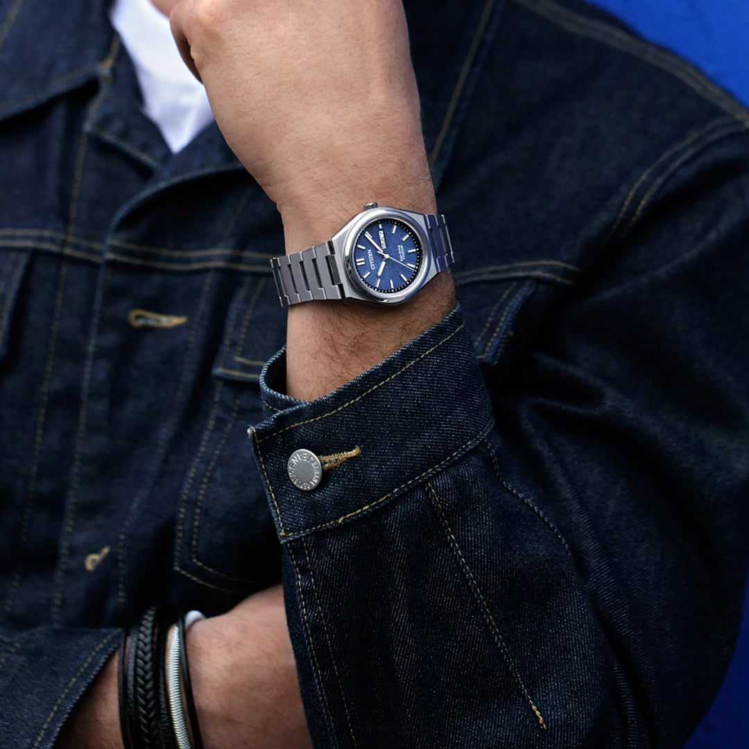 A person in a dark denim jacket rests one arm across their chest, wearing the CITIZEN Zenshin Three-hand Eco-Drive 39mm Watch by CITIZEN, featuring a Super Titanium™ case, blue face, and metal band.