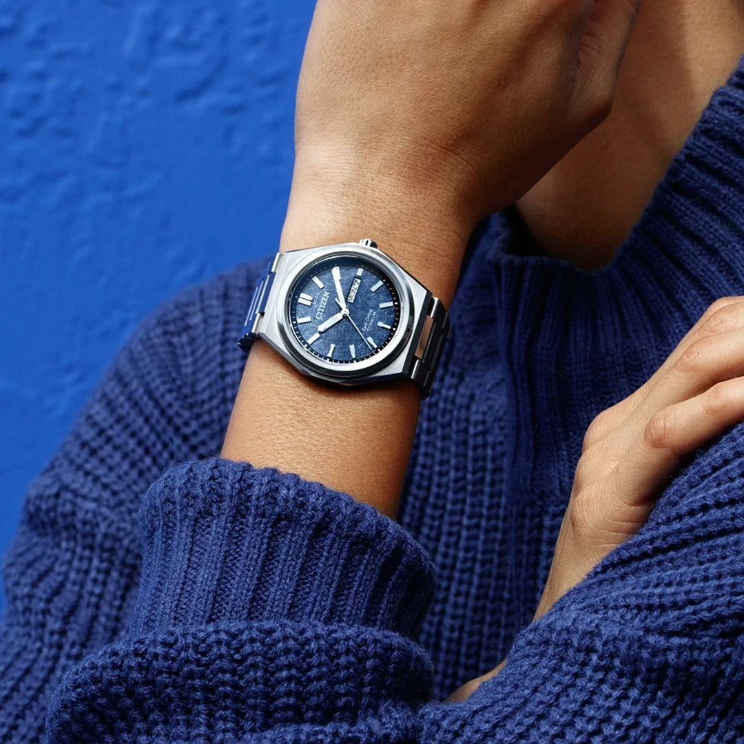 A person in a blue textured sweater rests their chin on their hand, highlighting the CITIZEN Zenshin Three-hand Eco-Drive 39mm Watch with a blue dial and silver case against a matching blue background.