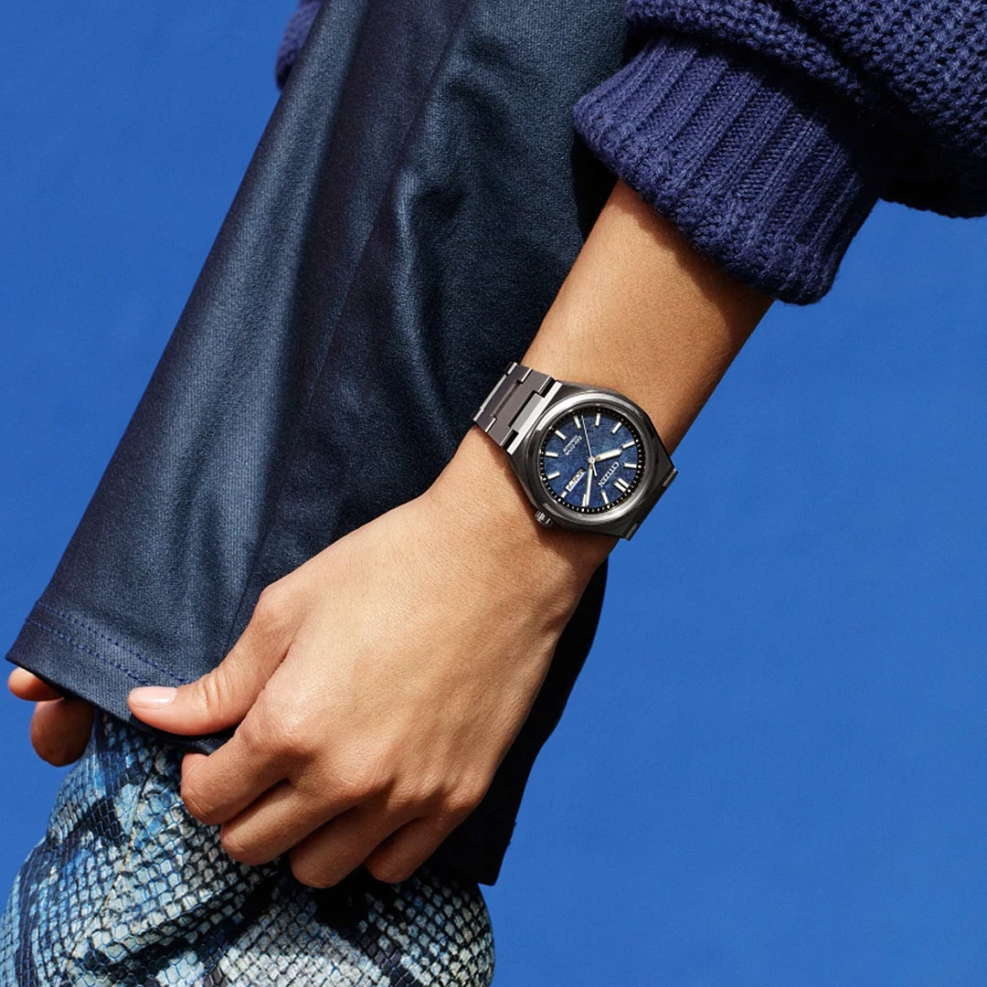 A person in a dark blue sweater and snakeskin-patterned pants displays the CITIZEN Zenshin Three-hand Eco-Drive 39mm Watch—a silver, water resistant timepiece with a blue face—against a solid blue background.