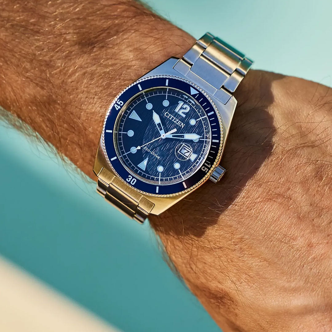 A close-up of a wrist wearing the CITIZEN Terra Force Eco-Drive 43mm Watch, featuring a blue dial and bezel, silver hour markers, and a metal bracelet by CITIZEN, set against a blurred aqua blue background.