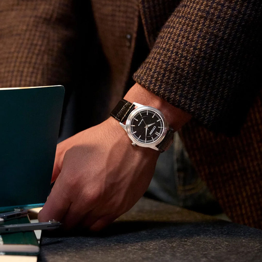 A person wearing a brown checkered jacket and a CITIZEN Rolan Eco-Drive 41mm Watch with a black dial and stainless steel case rests their hand beside a teal laptop on a desk.