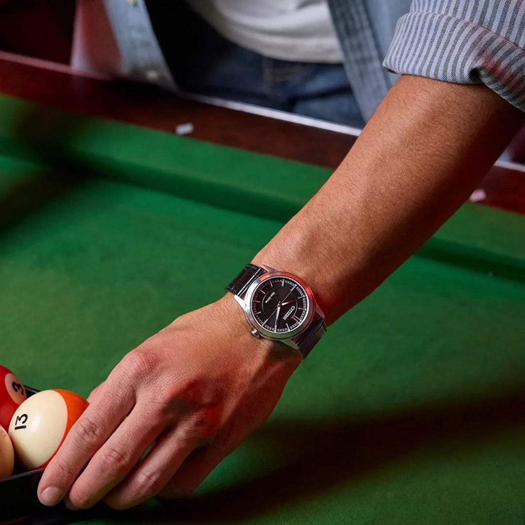 A person dressed in a rolled-up striped shirt reaches for billiard balls numbered 13 and 9 on a green pool table while wearing the CITIZEN Rolan Eco-Drive 41mm Watch, which features a sleek silver stainless steel case.