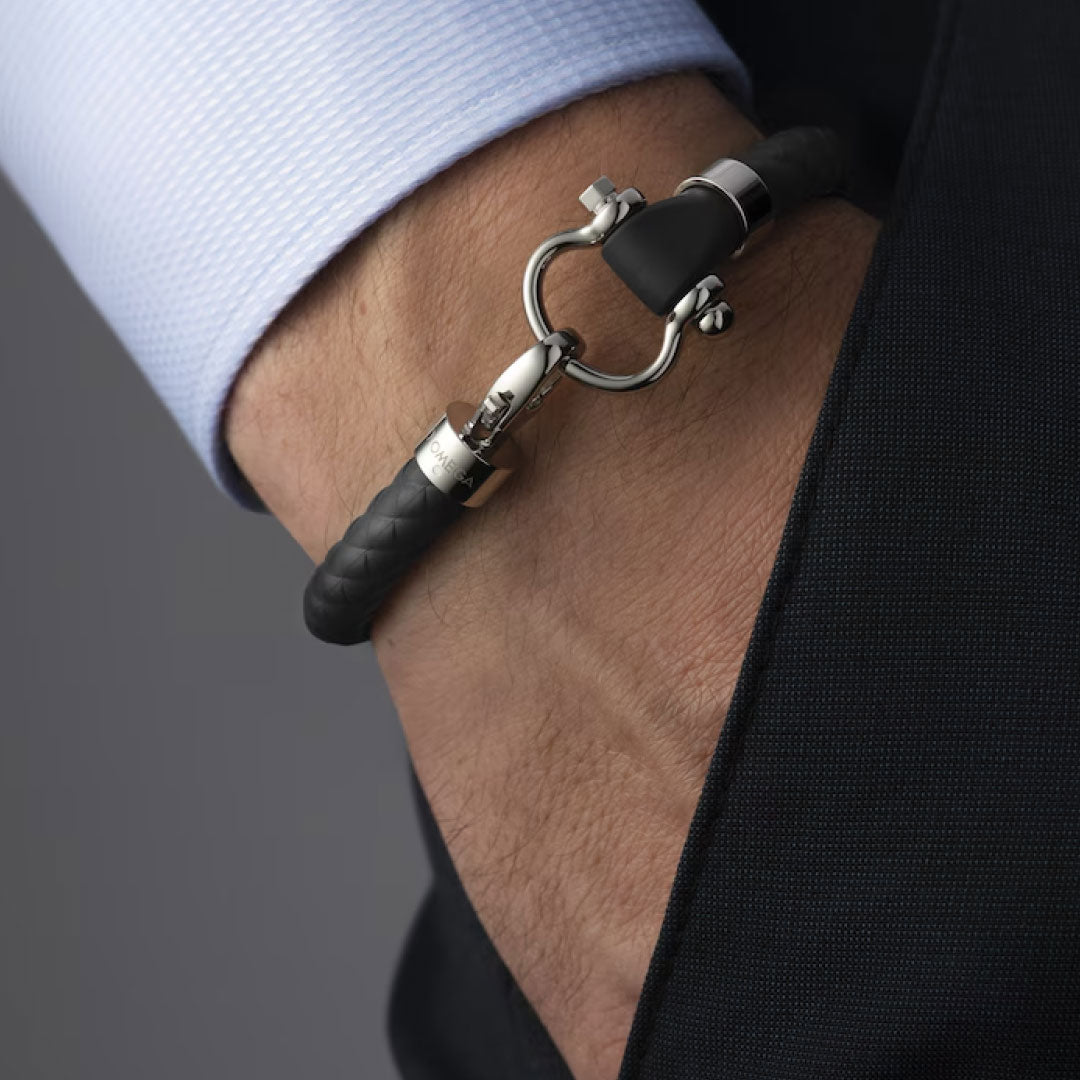 A close-up of a person's wrist wearing the OMEGA Aqua Sailing Rubber Bracelet in black and silver. The person is dressed in a light blue dress shirt and dark suit, with their hand in their pocket.