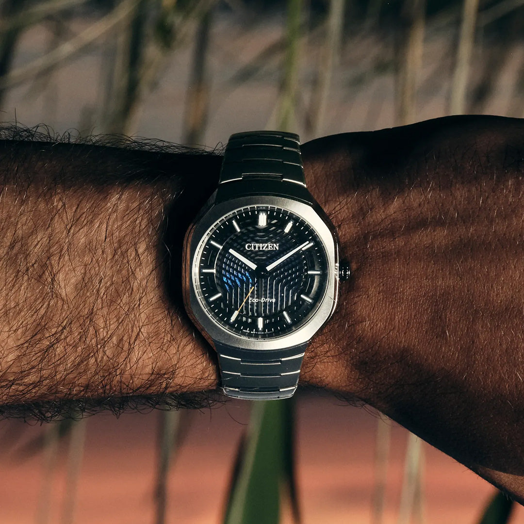 A CITIZEN Photon Eco-Drive 39.6mm Watch Limited Edition of 5,000 Pieces, crafted in stainless steel with a black patterned dial, adorns a person’s wrist. The watch face displays the time as 10:09, radiating an effortless sport-luxury appeal. A softly blurred outdoor background imparts a warm, natural ambiance to the scene.